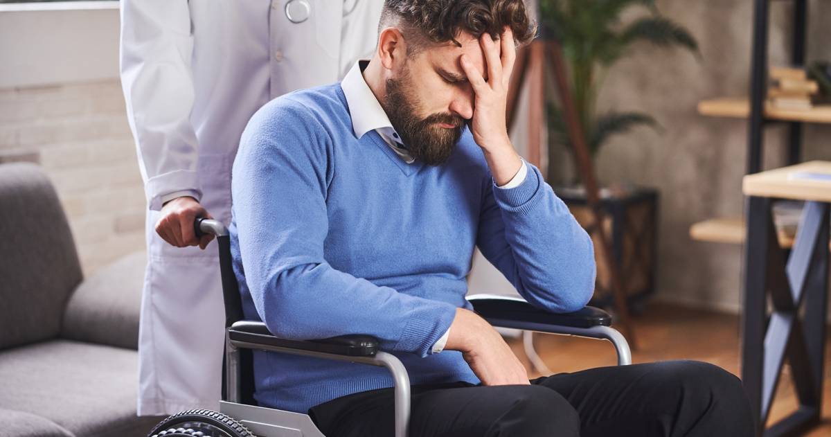 Trieste man in een rolstoel die een hand voor zijn gezicht houdt en geduwd wordt door een arts