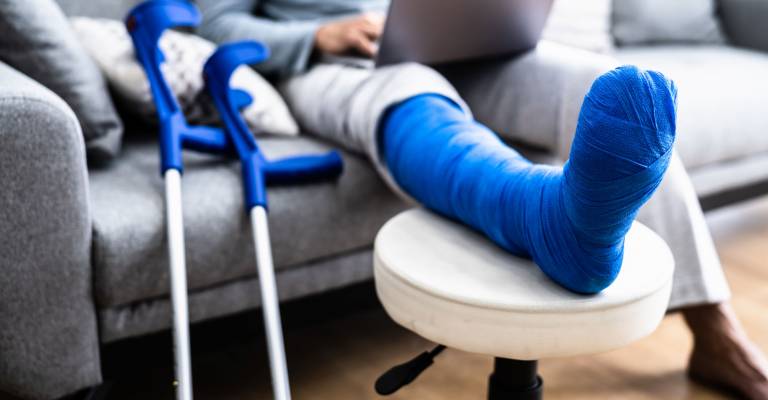 Een man met een gebroken been in blauw gips zittend op de bank met zijn been omhoog en krukken naast de bank